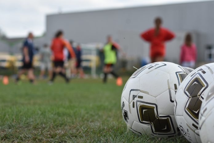 Image avec des ballons de foot en premier plan sur un terrain de football en herbe et en font on aperçoit les silhouettes floutées de footballeuses en entraînement